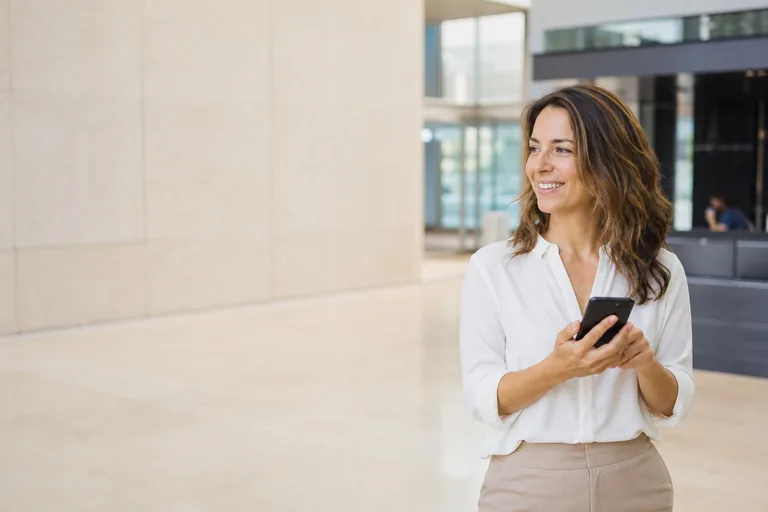 A woman in a bright office lobby holding her phone at chest height, relaxed.