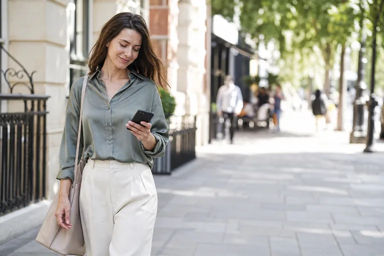 A woman on a city sidewalk glancing down at the phone in her hand, calm and starting to smile.